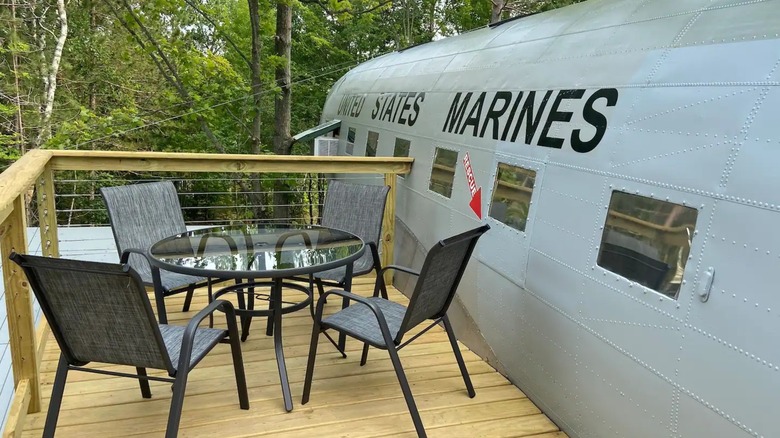 The side of a silver plane with attached deck and table with chairs.